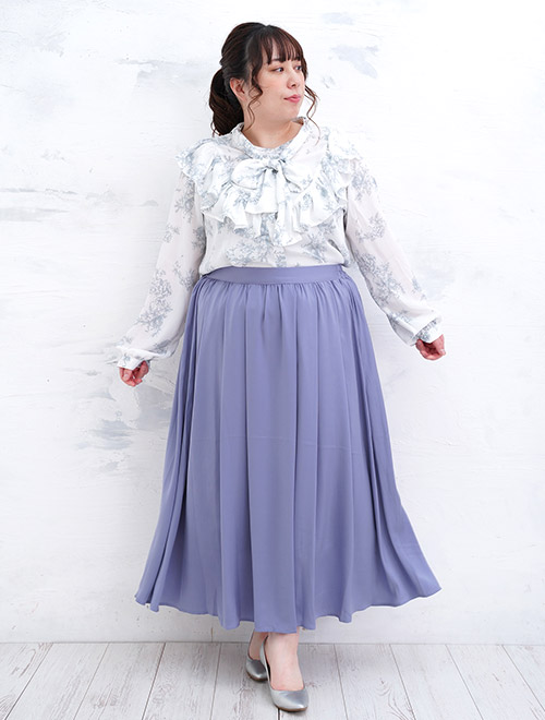 白地×ライトグレーの花柄リボンタイブラウスを着て、パープルブルーのAラインスカートを履く女性