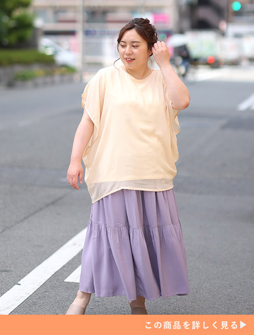シフォン素材を重ねたエクリュ色のトップスを着て、薄紫色のスカートを履く女性