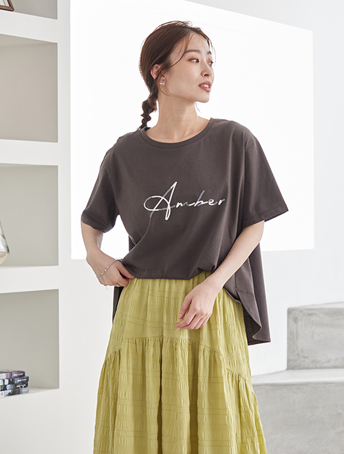チャコール色のロゴTシャツを、くすみイエローのスカートに前だけトップスインして着る女性