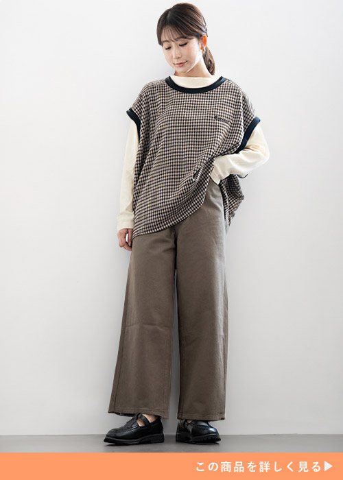 ホワイトトップスと格子柄ベストのレイヤードトップスにブラウンパンツを履く女性